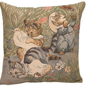 Puede incluir: Una almohada de tapiz con tres gatitos en un entorno de jardín. Los gatitos llevan ropa y están rodeados de flores y vegetación. La almohada está hecha de un tejido suave y tejido con un brillo sutil.