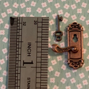 May include: A miniature door knob and key set in a copper finish. The knob has a decorative design and a lever handle. The key is a simple design with a round hole. The set is measured against a ruler, showing it is approximately 1 inch in size.