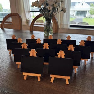 May include: A collection of small black chalkboards on wooden easels. The easels are made of light-colored wood. A vase of white flowers sits in the background, near a window.