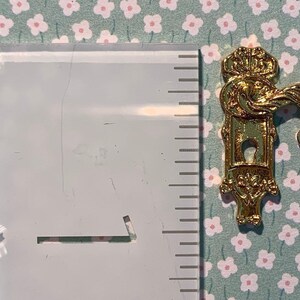May include: Gold-toned metal door knob with a decorative keyhole and a key. The knob has a decorative design with a crown on top and a bird-like handle. The key is a simple design with a round head.