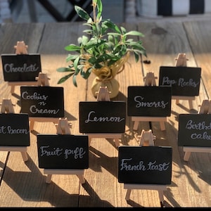May include: Ten small black chalkboard signs with white lettering on wooden easels. The signs are arranged on a wooden surface and feature various food and dessert names, including "Chocolate", "Cookies n Cream", "Sweet Bacon", "Lemon", "Fruit Puff", "Pop Rocks", "Smores", "French Toast", and "Birthday Cake".