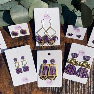 May include: Assortment of purple and gold earrings with various geometric shapes and designs. The earrings are displayed on white cards with the word "snella" printed on them.