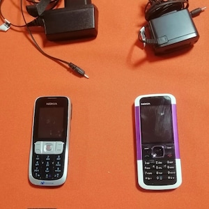 May include: Two Nokia mobile phones with physical keypads, two black wall chargers with coiled cords, and two rectangular lithium-ion batteries labeled BL-4B and BL-4BA. The devices are arranged in pairs, featuring black and silver or purple accents.
