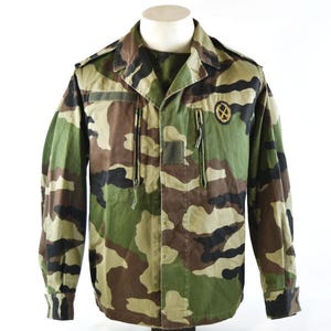 May include: A long-sleeve military-style jacket featuring a camouflage pattern in shades of green, brown, and black. The jacket includes a fold-down collar, vertical zippered chest pockets, shoulder epaulets, and a circular patch on the left chest.