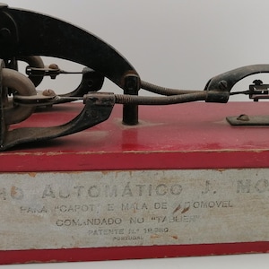 May include: A vintage car hood latch mechanism in a red wooden box. The mechanism is made of metal and has a silver knob. The box has a label that reads "FECHO AUTOMÁTICO J. MOTA PARA CAPOT, E MIRA DE AUTOMOVEL COMANDADO NO 414.125 PATENTE IL 14.330".