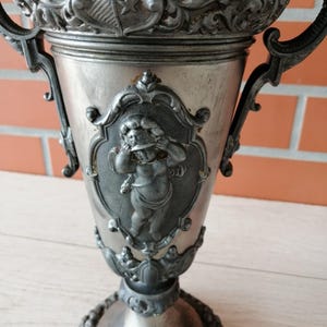 May include: An antique silver-colored chalice with ornate detailing. The cup features a cherub design, decorative handles, and a detailed base. The top rim has a shield design with stars. The chalice is likely made of metal.