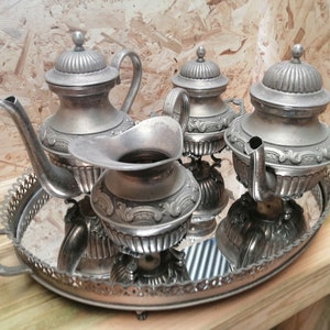 Op de afbeelding: Een zilveren theeservies met een theepot, roomkan, suikerpot en een dienblad met een gespiegelde oppervlak. De set heeft sierlijke details en een klassiek ontwerp.