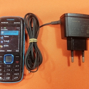 May include: A black Nokia XpressMusic mobile phone with a blue trim, a numeric keypad, and a screen displaying a messaging menu, connected to a black wall charger with a two-prong plug by a long, coiled black power cord.