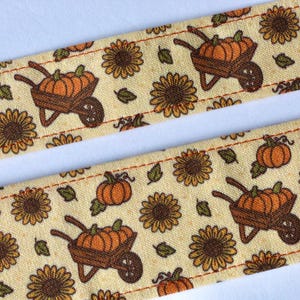 May include: Close-up of two strips of fabric with a repeating pattern of pumpkins in wheelbarrows, sunflowers, and leaves on a cream background. The fabric is stitched with orange thread, creating a border.