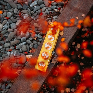 May include: Yellow keychain with a repeating moon phase design. The keychain is lying on a wooden surface with a background of pebbles and autumn leaves. The moon phases are orange and white.
