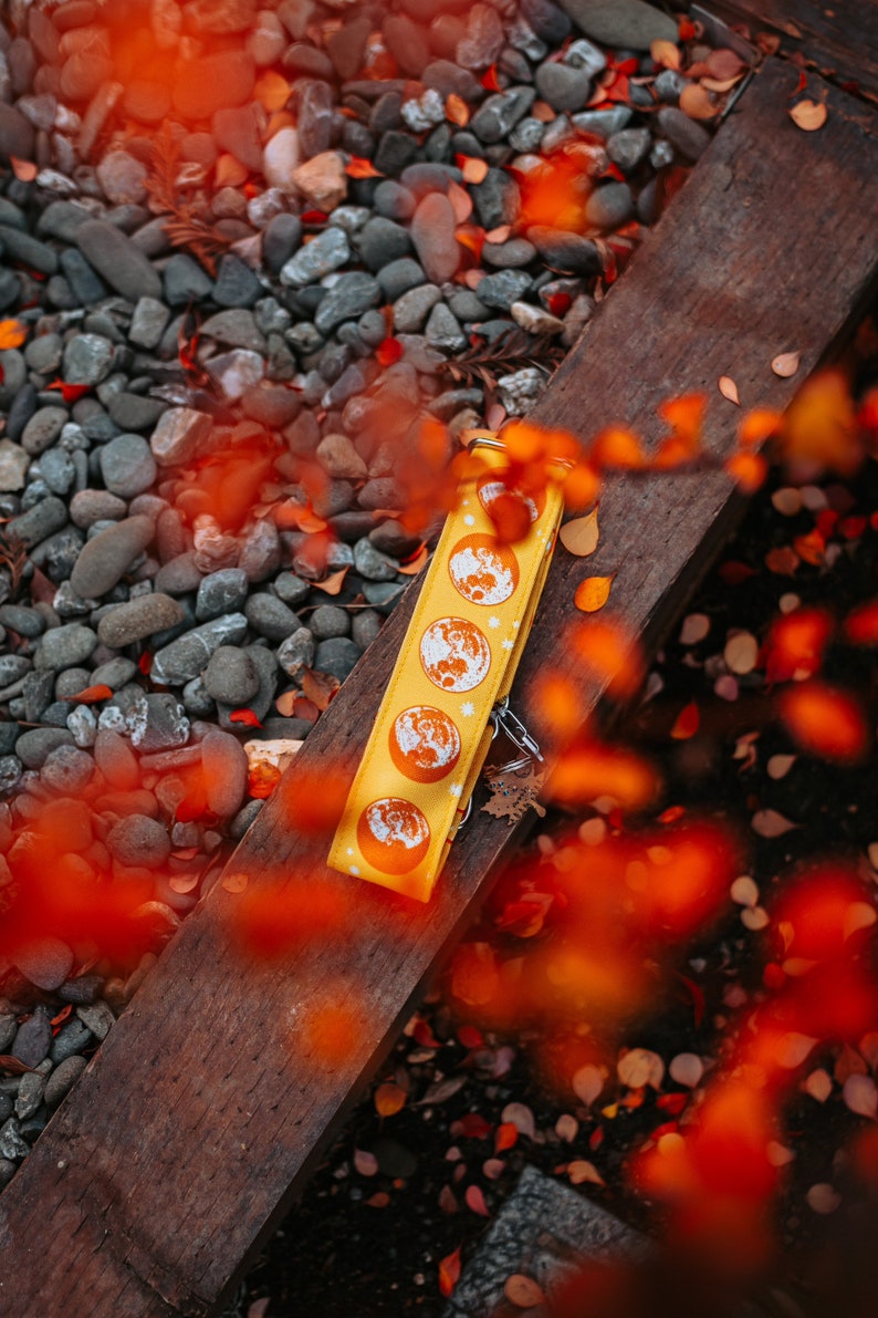 May include: Yellow keychain with a repeating moon phase design. The keychain is lying on a wooden surface with a background of pebbles and autumn leaves. The moon phases are orange and white.