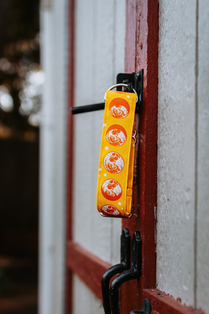 May include: A yellow fabric keychain with a repeating pattern of orange moons and white stars. The keychain is hanging on a red and white wooden door with black hardware.