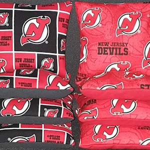 May include: Set of six bean bags featuring the New Jersey Devils logo. The bean bags are red and black with a white outline. The logo is a red and white stylized 'N' with a devil's tail.