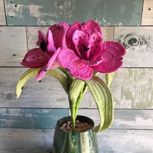 May include: A crocheted pink flower bouquet in a green and gold marbled vase. The vase is sitting on a black and pink table.