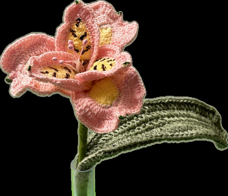 May include: A crocheted pink flower with yellow accents and a green crocheted stem in a clear glass vase.