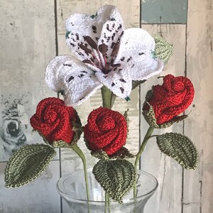 May include: A bouquet of crocheted flowers in a clear glass vase. The bouquet includes three red roses and one white lily.