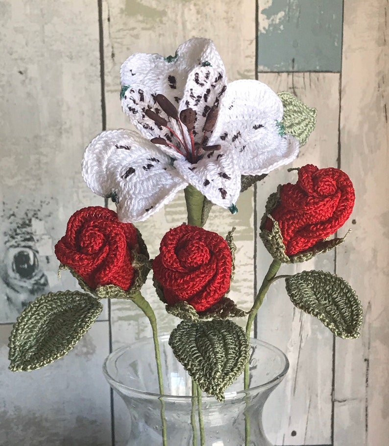 May include: A bouquet of crocheted flowers in a clear glass vase. The bouquet includes three red roses and one white lily.
