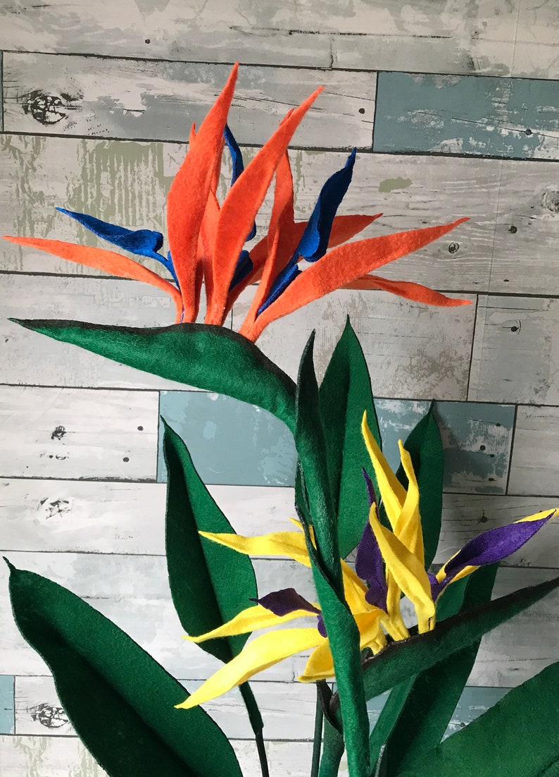 Two Bird of Paradise felt flowers. One flower with orange tepals and blue petals, and the second flower with yellow tepals and purple petals surrounded by 7 leaves. 3 small leaves, 2 medium leaves, and 2 large leaves.