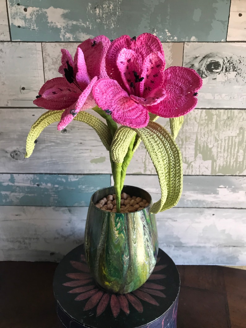 May include: A crocheted pink flower bouquet in a green and gold marbled vase. The vase is sitting on a black and pink table.