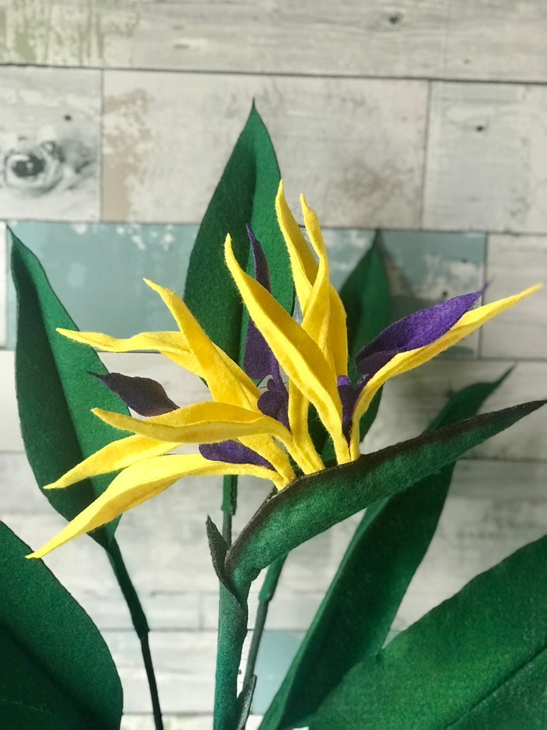 Bird of Paradise felt flowers with yellow tepals and purple petals surrounded by 7 leaves. 3 small leaves, 2 medium leaves, and 2 large leaves.