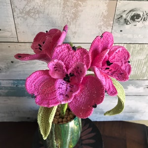 May include: A crocheted bouquet of pink flowers in a green and gold vase. The flowers have black centers and green leaves.