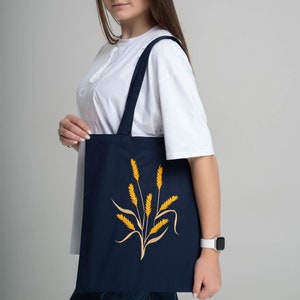 May include: Navy blue tote bag featuring a golden wheat stalk design. The bag has long straps for shoulder or hand carry. The wheat design is centered on the front of the bag. The bag is made of a sturdy material.