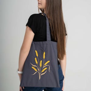 May include: A woman is carrying a gray tote bag with a yellow wheat design. The bag has long straps and is made of a canvas material.