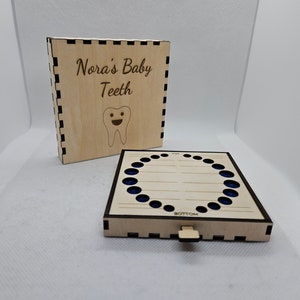 May include: A wooden box with a lid that opens to reveal a compartment for storing baby teeth. The lid is engraved with the text "Nora's Baby Teeth" and a smiling cartoon tooth. The box has a dark brown border and a light wood finish.
