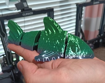 May include: A 3D printed green and blue shark figurine with a textured surface. The figurine is being held in a person's hand.
