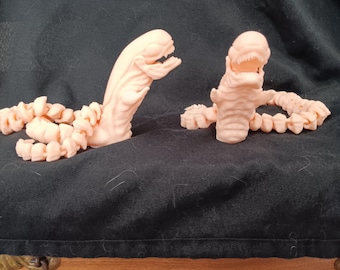 May include: Two light pink, worm-like sculptures with detailed, open-mouthed heads and segmented bodies. One is upright, the other is horizontal. The sculptures appear to be 3D-printed and are set against a dark background.