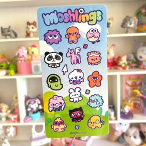 Op de afbeelding: Een rechthoekig stickervel met de tekst Moshlings bovenaan en een verzameling kleurrijke stripfiguren, waaronder een panda, een eenhoorn, een geest en diverse wezens in paars, oranje, roze, groen en blauw op een lichtblauwe achtergrond.