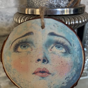 May include: A circular wooden ornament depicting a celestial face with blue and pink hues, long eyelashes, and glitter. The ornament has a natural wood edge and a jute string for hanging. The design has a whimsical, vintage aesthetic.