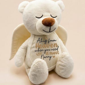 May include: A cream-colored plush teddy bear with soft fur, closed eyes, and a brown nose. The bear features attached angel wings on its back and embroidered text on its chest that reads: A hug from Heaven, when you need it most, Nanny.