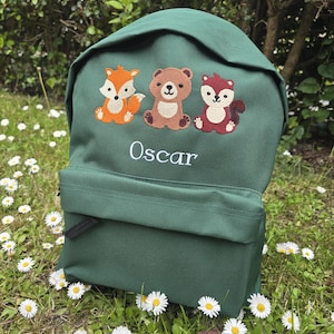 May include: A dark green backpack with a front pocket and embroidered animal designs: a fox, a bear, and a squirrel. The name "Oscar" is embroidered below the animals. The backpack is on a grassy surface with daisies.