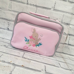 May include: Pink lunch bag with a grey trim. The bag features an embroidered design of two bunnies with flowers and leaves. The name "Olivia" is embroidered below the bunnies.
