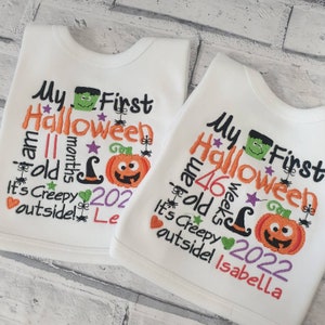 May include: Two white baby onesies with embroidered Halloween themed text. The text on the onesies reads "My First Halloween I am 11 months old It's Creepy outside! Le" and "My First Halloween I am 46 weeks old It's Creepy outside! Isabella".