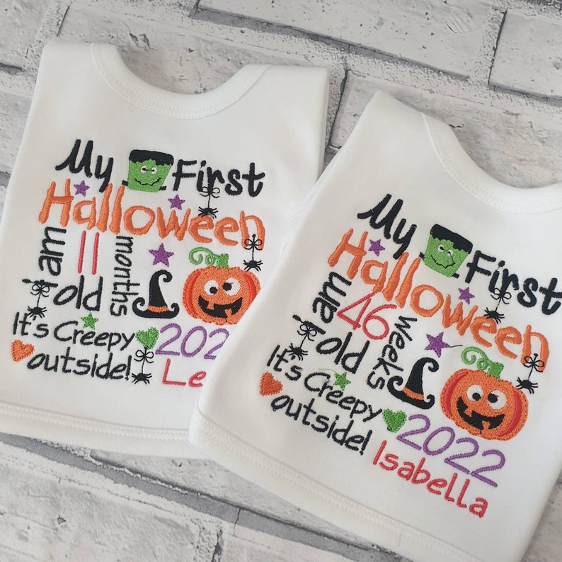 May include: Two white baby onesies with embroidered Halloween themed text. The text on the onesies reads "My First Halloween I am 11 months old It's Creepy outside! Le" and "My First Halloween I am 46 weeks old It's Creepy outside! Isabella".