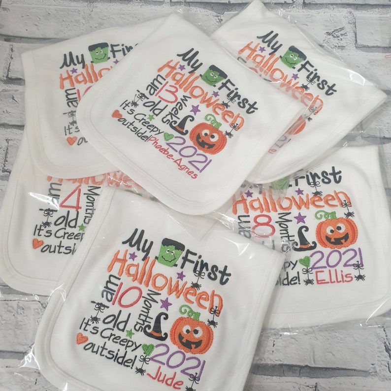 May include: A stack of white baby bibs with embroidered text that says "My First Halloween" and includes a pumpkin, witch hat, and stars. The bibs are personalized with different names and ages.