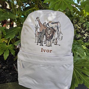 Op de afbeelding: Een lichtgrijze rugzak met een geborduurd ontwerp van verschillende safaridieren, waaronder een giraffe, een zebra, een leeuw en een olifant. De naam "Ivor" is eronder geborduurd. De rugzak heeft een voorvak met rits en verstelbare bandjes.