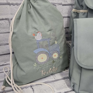 May include: A sage green drawstring backpack featuring an embroidered blue tractor with a small bird perched on top. The name Teddy is embroidered in light brown script below the tractor. The bag has thick white rope straps for closure.