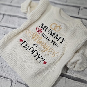 May include: A cream-coloured knit jumper featuring embroidered text that reads MUMMY WILL YOU Marry MY DADDY? The design includes two gold rings, small red hearts, and decorative gold swirls stitched onto the front of the ribbed fabric.