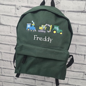May include: A dark green backpack with black straps and a front pocket. The backpack features embroidered images of a blue train, a yellow excavator, and a green tractor. The name "Freddy" is embroidered in white.