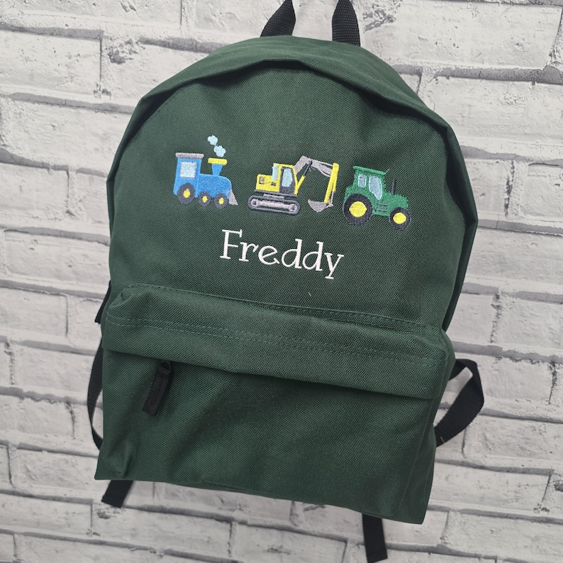 May include: A dark green backpack with black straps and a front pocket. The backpack features embroidered images of a blue train, a yellow excavator, and a green tractor. The name "Freddy" is embroidered in white.