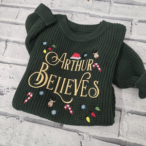 May include: A dark green long-sleeve knit jumper featuring gold embroidered text that reads Arthur Believes. The design includes small embroidered accents of a red Santa hat, reindeer faces, candy canes, blue snowflakes, and colourful light bulbs.