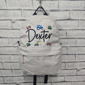 May include: A pale grey backpack featuring embroidered construction vehicles in various colours and the name "Dexter" in black script. It has a front pocket, a top handle, and black shoulder straps.