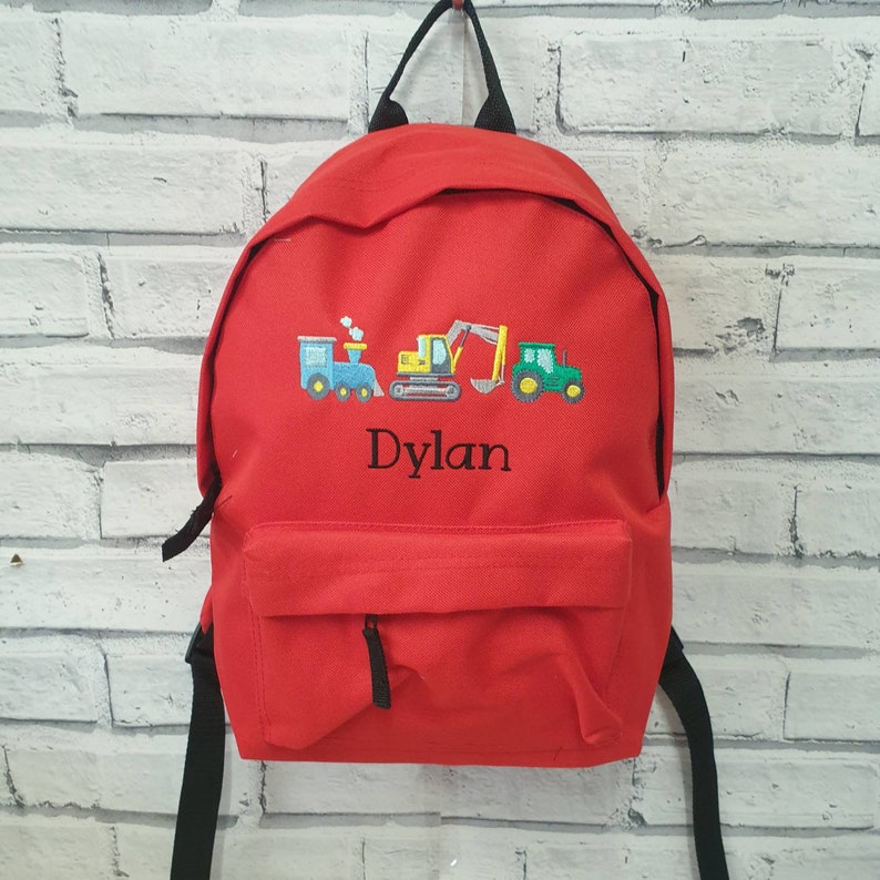 May include: A red backpack with a front pocket and black straps. The backpack features an embroidered design of a blue train, a yellow excavator, and a green tractor. The name "Dylan" is embroidered below the vehicles.
