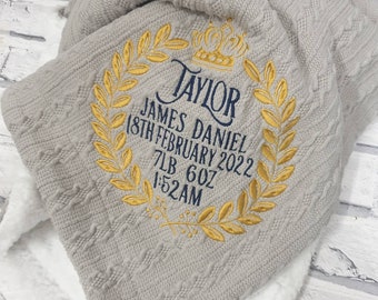 May include: A gray knitted baby blanket with a gold embroidered crown and laurel wreath. The blanket is personalized with the name "Taylor James Daniel", the date "18th February 2022", the weight "7lb 6oz", and the time "1:52AM".