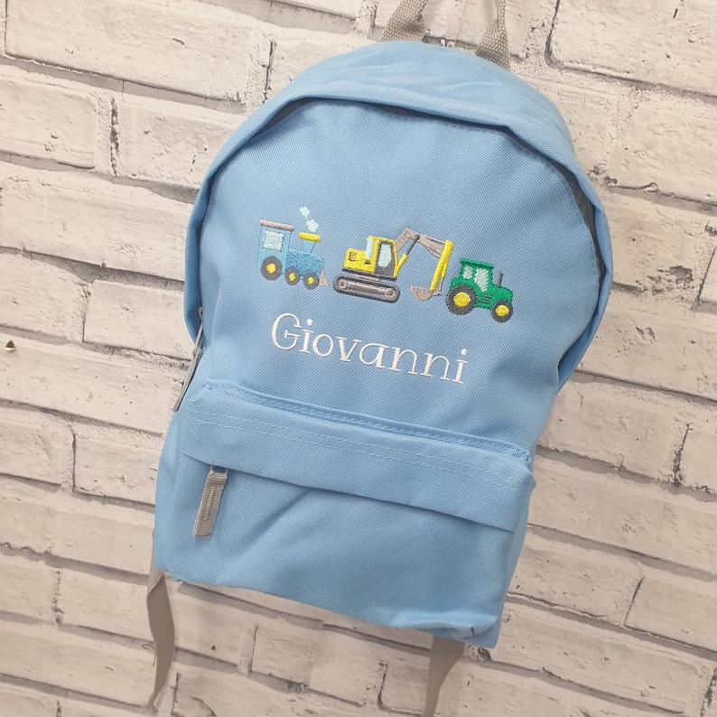 May include: Light blue backpack with embroidered train, excavator, and tractor designs. The name "Giovanni" is embroidered below the vehicles. The backpack has a front pocket and gray straps.