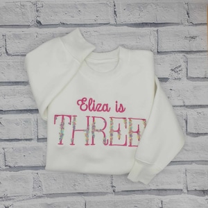 Peut inclure: Un sweat-shirt blanc avec l'inscription brodée "Eliza is THREE" en rose et lettres fleuries. Le sweat-shirt est plié sur un fond de mur en briques blanches.