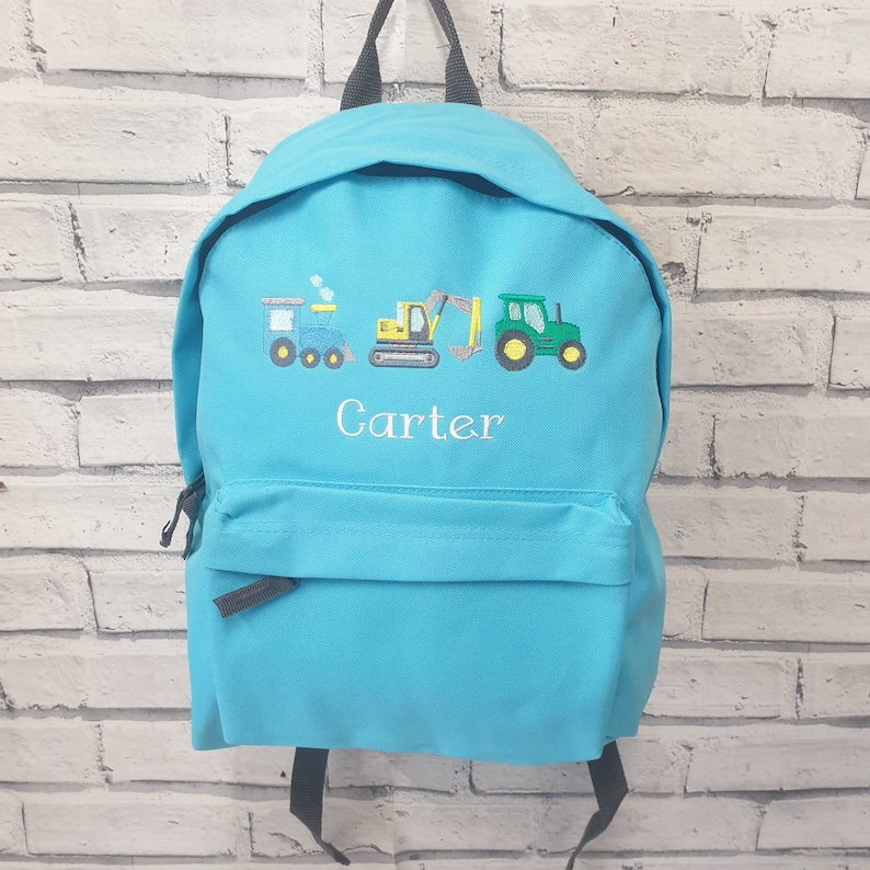 May include: A turquoise backpack with a front pocket and black straps. The backpack features embroidered images of a blue train, a yellow excavator, and a green tractor. The name "Carter" is embroidered in white below the vehicles.